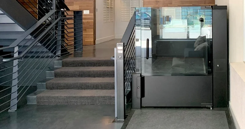Indoor vertical platform lift with glass enclosure installed beside a staircase, providing wheelchair‑accessible access in a modern commercial interior.