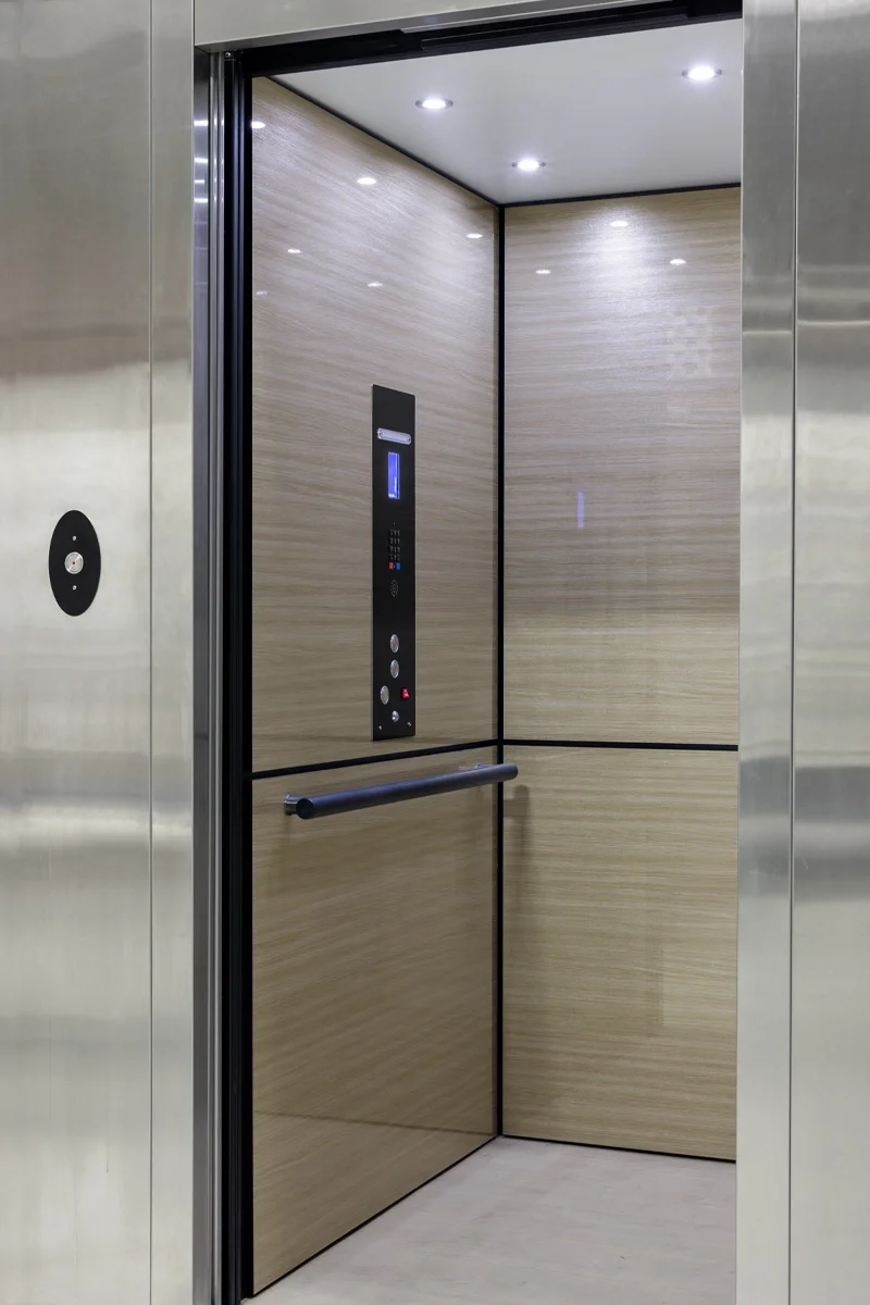 Modern residential elevator interior with stainless steel doors, light wood‑grain wall panels, illuminated control panel, safety handrail, and recessed LED ceiling lights.