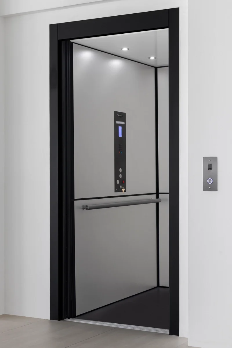 Modern home elevator with black frame and open door, featuring a gray interior, stainless steel control panel, horizontal handrail, recessed ceiling lights, and wall-mounted call button in a residential hallway.