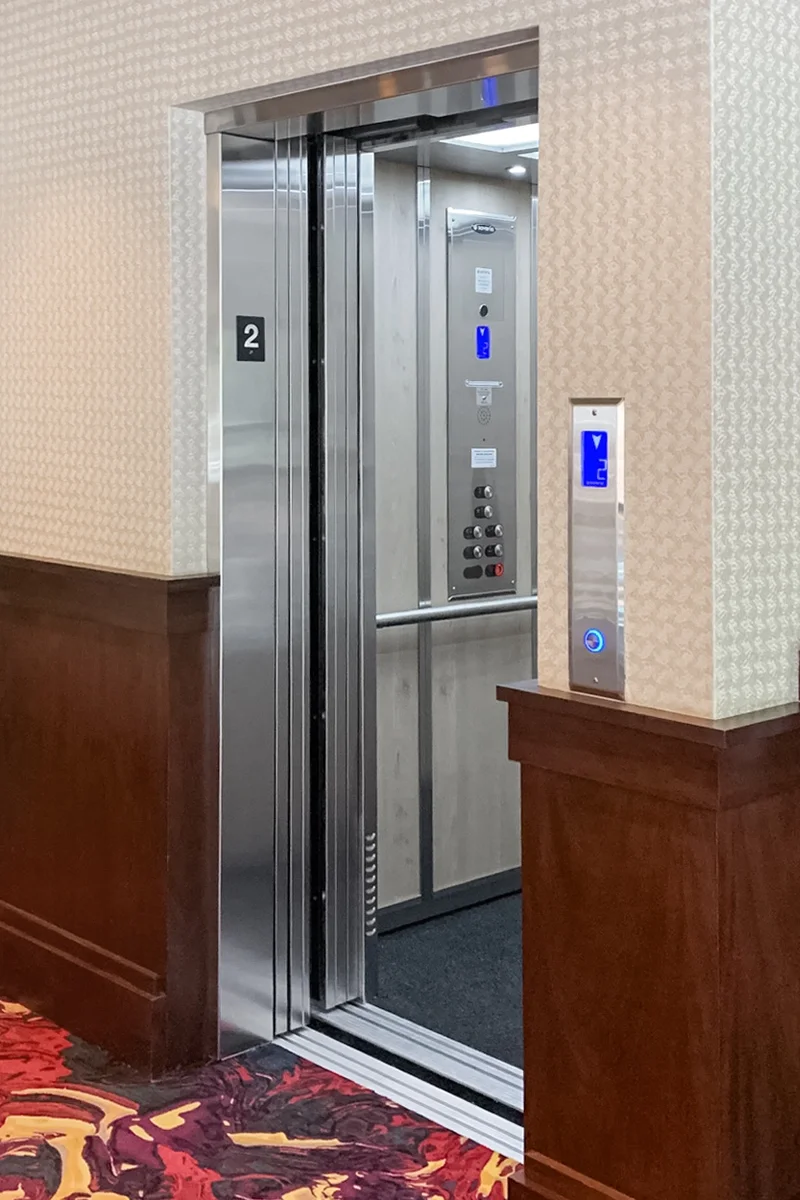 Commercial elevator with stainless steel doors, illuminated interior control panel, floor indicator, and wall‑mounted call button installed in a hotel or commercial hallway.