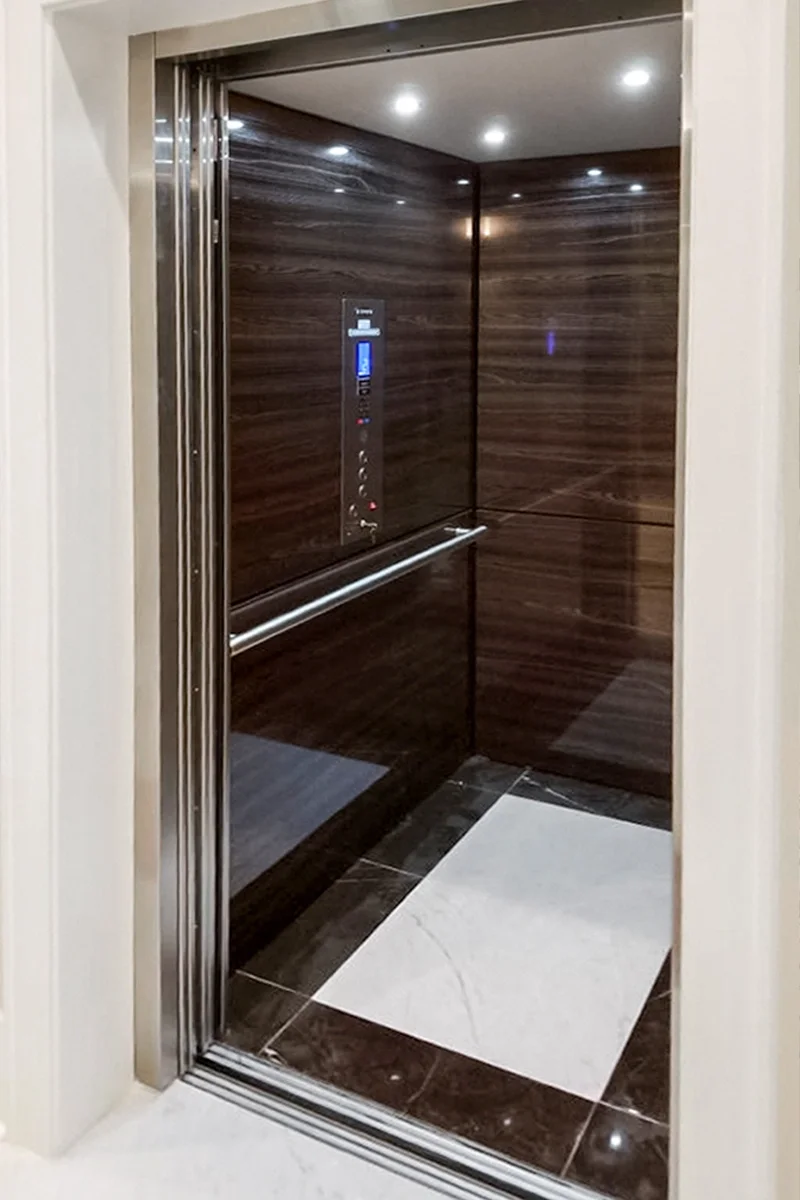 Luxury residential elevator interior with dark wood panel walls, stainless steel control panel, handrail, recessed lighting, and open doorway in a modern home.
