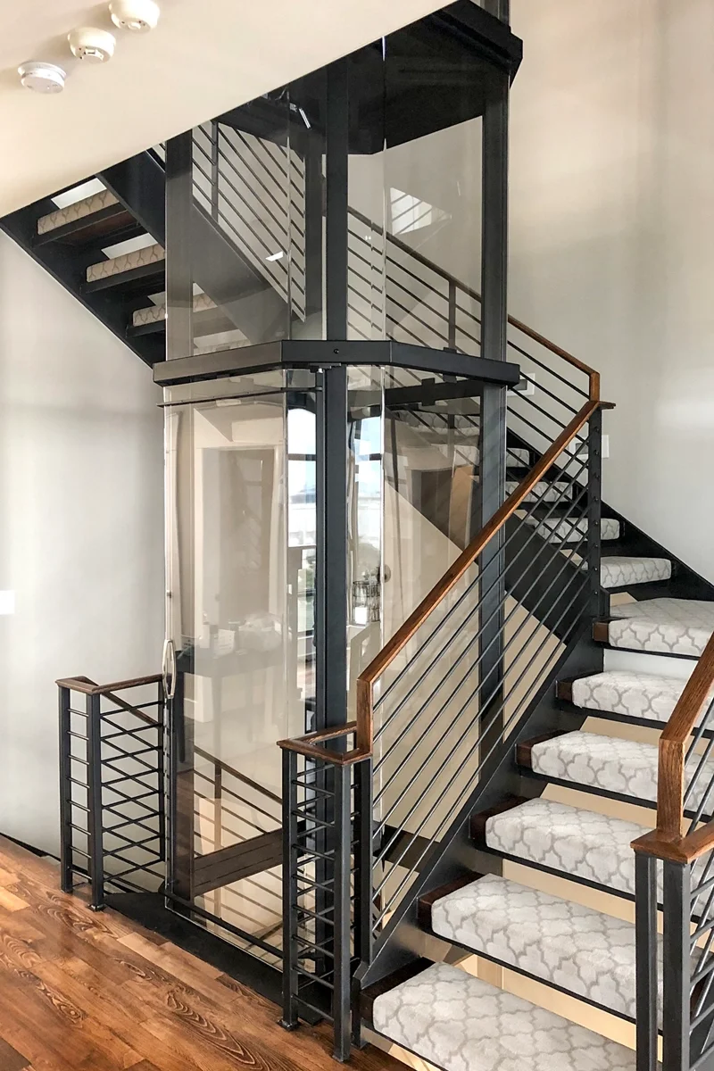 Panoramic glass residential elevator installed between staircases in a modern luxury home, featuring black frame design, open-concept layout, and contemporary railing.