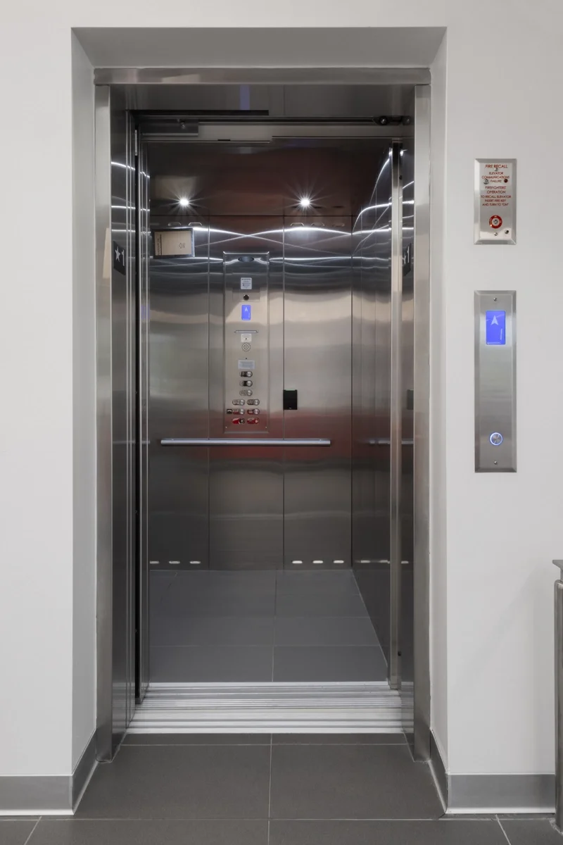 Commercial stainless steel elevator with open doors, illuminated control panel, call buttons, and modern interior installed in a professional office or commercial building.
