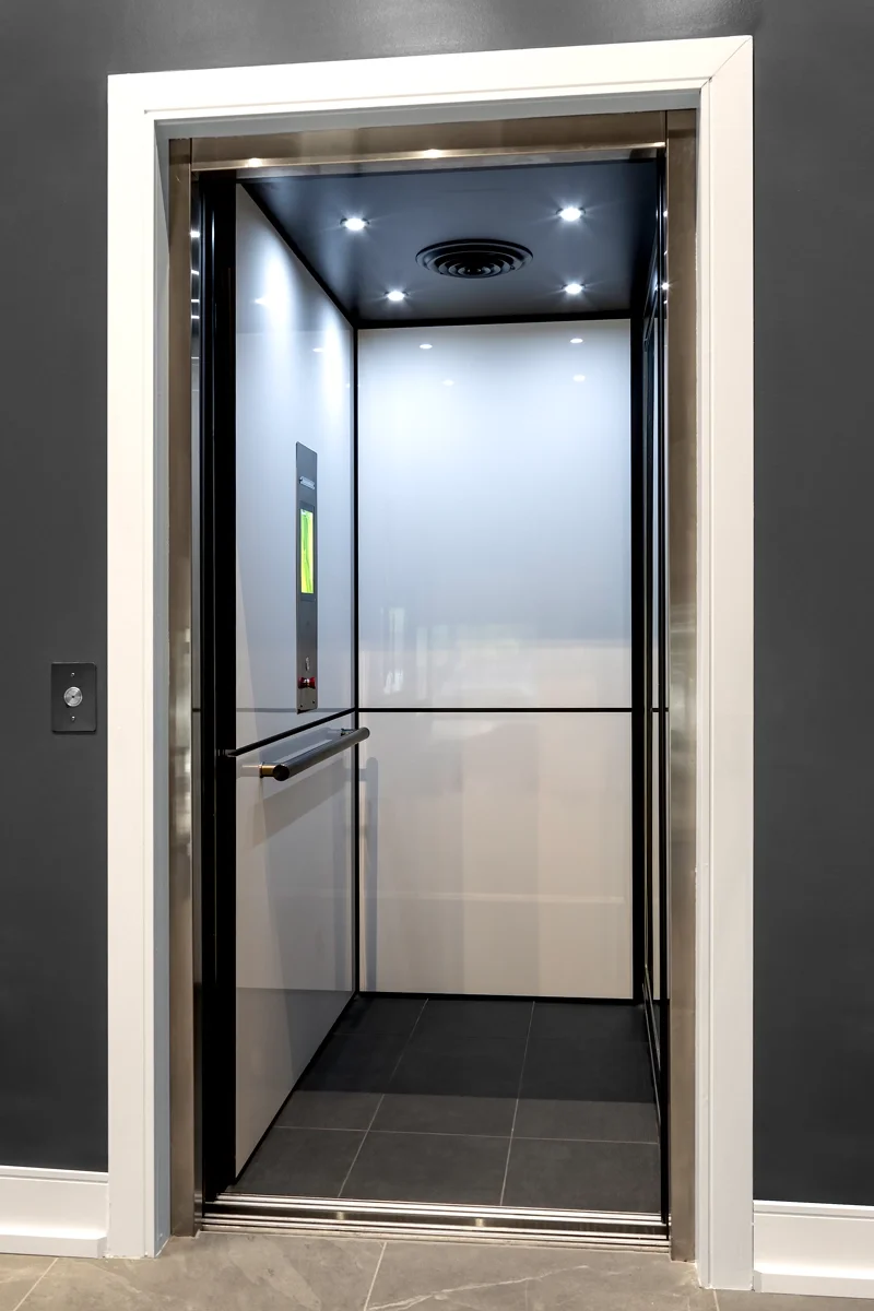 Modern home elevator with stainless steel trim, white interior walls, recessed LED lighting, handrail, and open door installed in a residential setting.