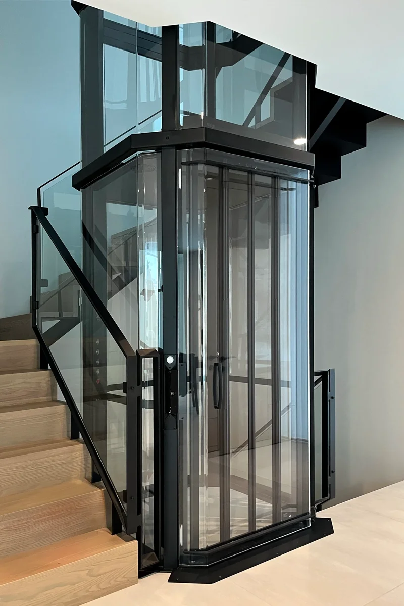 Panoramic glass residential elevator with black frame installed beside a modern staircase, providing stylish and accessible vertical mobility in a luxury home.