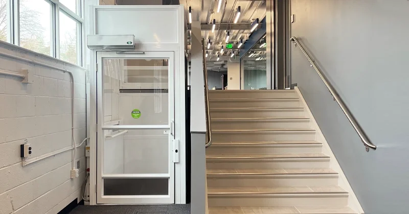 Indoor vertical platform lift with white enclosure and glass door installed next to a staircase in a modern commercial or office building for wheelchair accessibility.