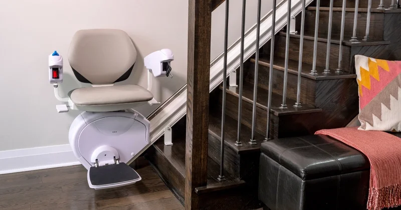 Curved residential stairlift with upholstered seat, armrests, footrest, and wall‑mounted control installed on a wooden staircase for home accessibility.