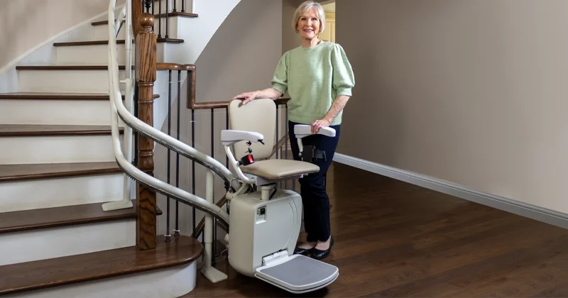 Twin rail stairlift with curved double rail track installed on a residential staircase, featuring an upholstered seat and an older woman standing beside it.