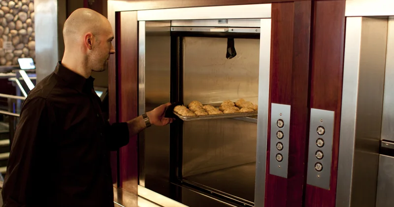 Restaurant material lift transporting food trays, with a staff member loading baked goods into a commercial service elevator.