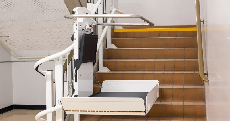 Inclined platform lift installed on an indoor staircase, featuring a fold‑down wheelchair platform, guide rails, and safety arms providing accessible mobility between floor levels in a commercial or institutional building.