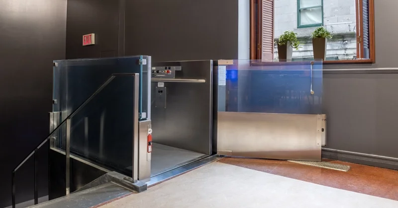 Indoor vertical platform lift with glass barriers and stainless steel enclosure, providing wheelchair accessibility between floor levels in a commercial building.