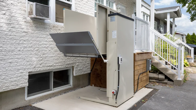 Outdoor vertical platform lift installed at a residential home entrance, featuring an enclosed accessibility lift providing wheelchair access to a raised porch.