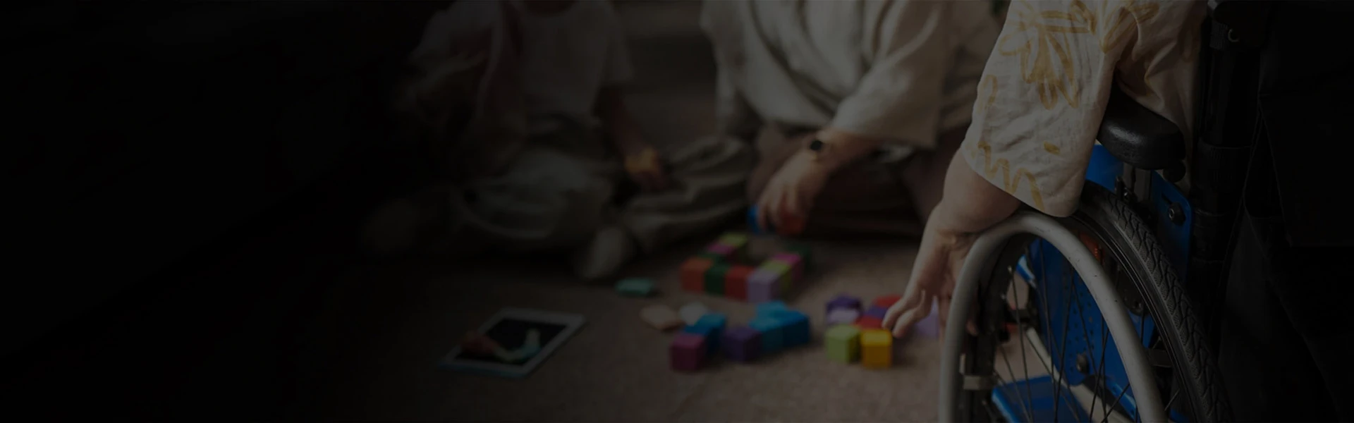 Wheelchair user reaching toward colorful toy blocks on the floor beside children playing.
