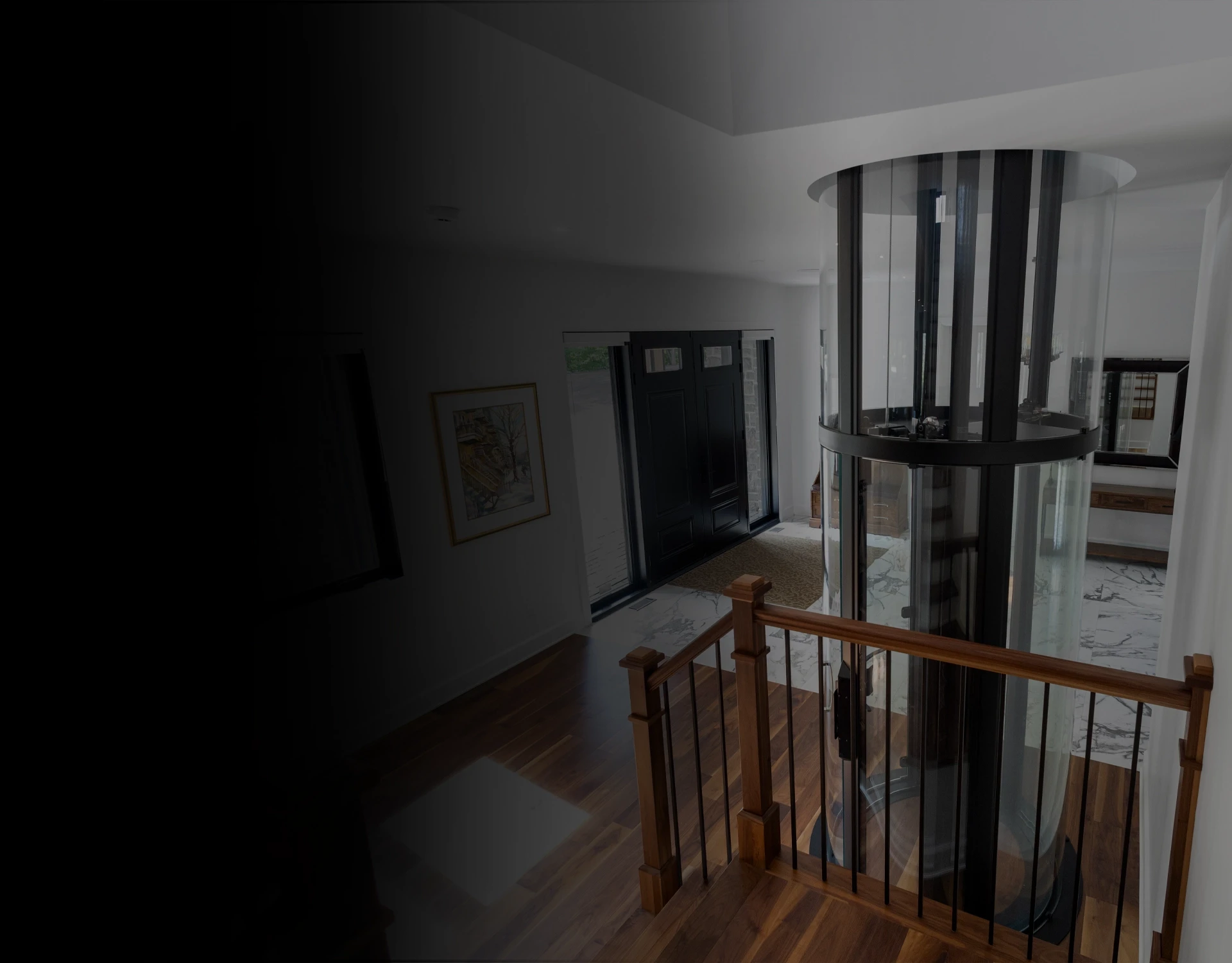Glass cylindrical residential elevator beside a wooden stair railing in a bright modern foyer