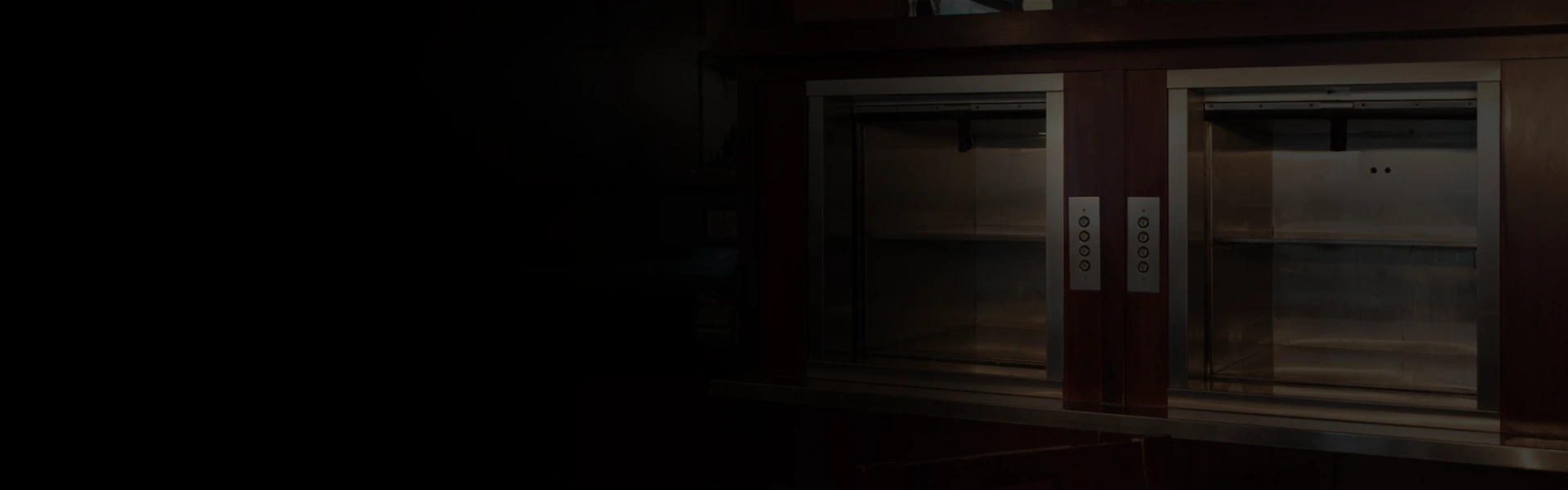 Dark wood-paneled dumbwaiter with stainless openings in a low-lit interior