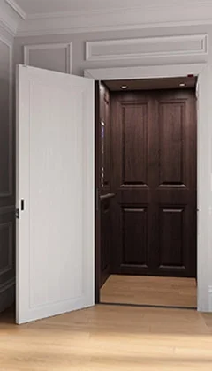 An open home elevator with white exterior doors and a dark wood-paneled interior, installed in a residential setting with light wood flooring and decorative wall panels.