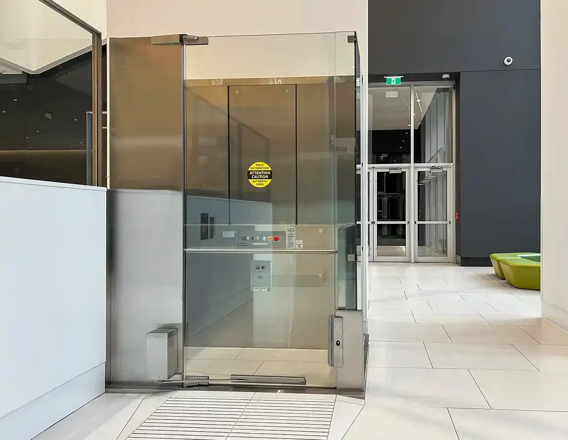 Glass-enclosed commercial elevator at an indoor landing with stainless steel framing and transparent doors