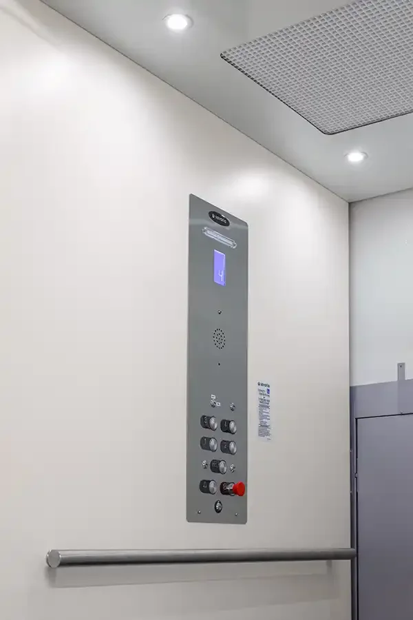Vertical platform lift interior showing wall-mounted control panel with floor indicator, emergency stop button, and handrail