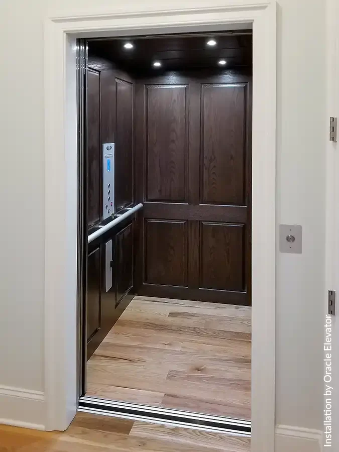Luxury residential elevator interior with dark wood panel walls, stainless steel handrail, recessed LED lighting, and open doorway in a modern home.