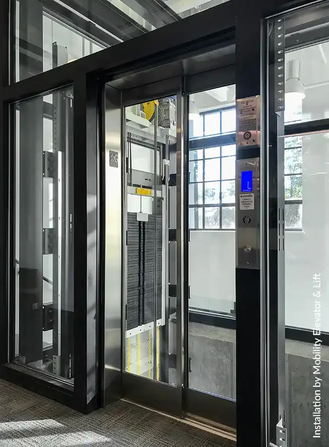 Glass enclosed commercial elevator with black metal frame and stainless steel doors installed inside a modern building, featuring illuminated control panel and transparent cab for accessible vertical transportation.