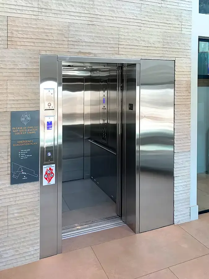 Stainless steel commercial elevator with open doors installed in a modern building lobby, featuring illuminated call buttons, control panel, and accessible entry for vertical mobility between floors.