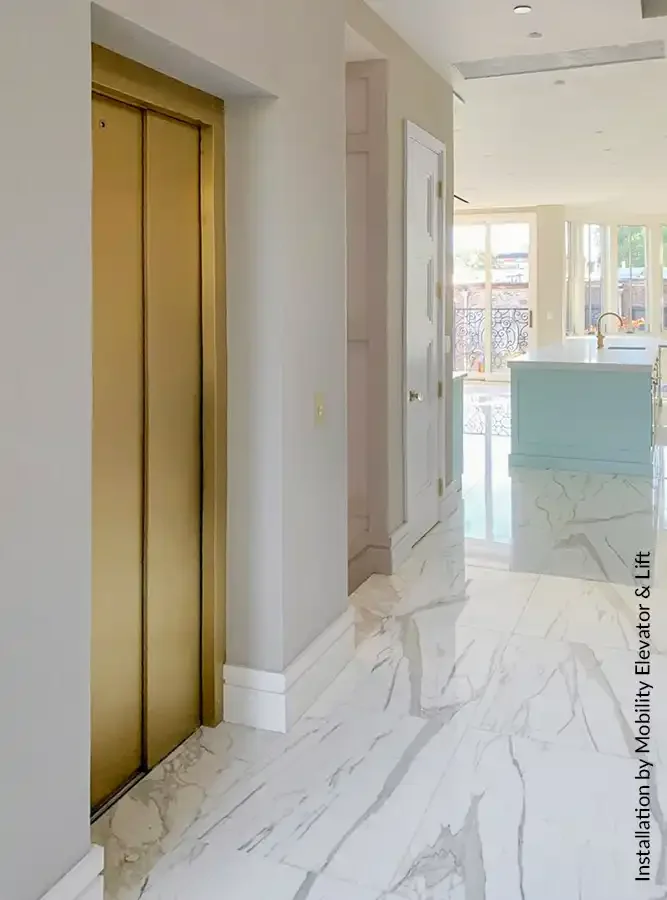 Residential elevator with brushed brass sliding doors installed in a luxury home, featuring marble-look flooring and modern open‑concept interior.