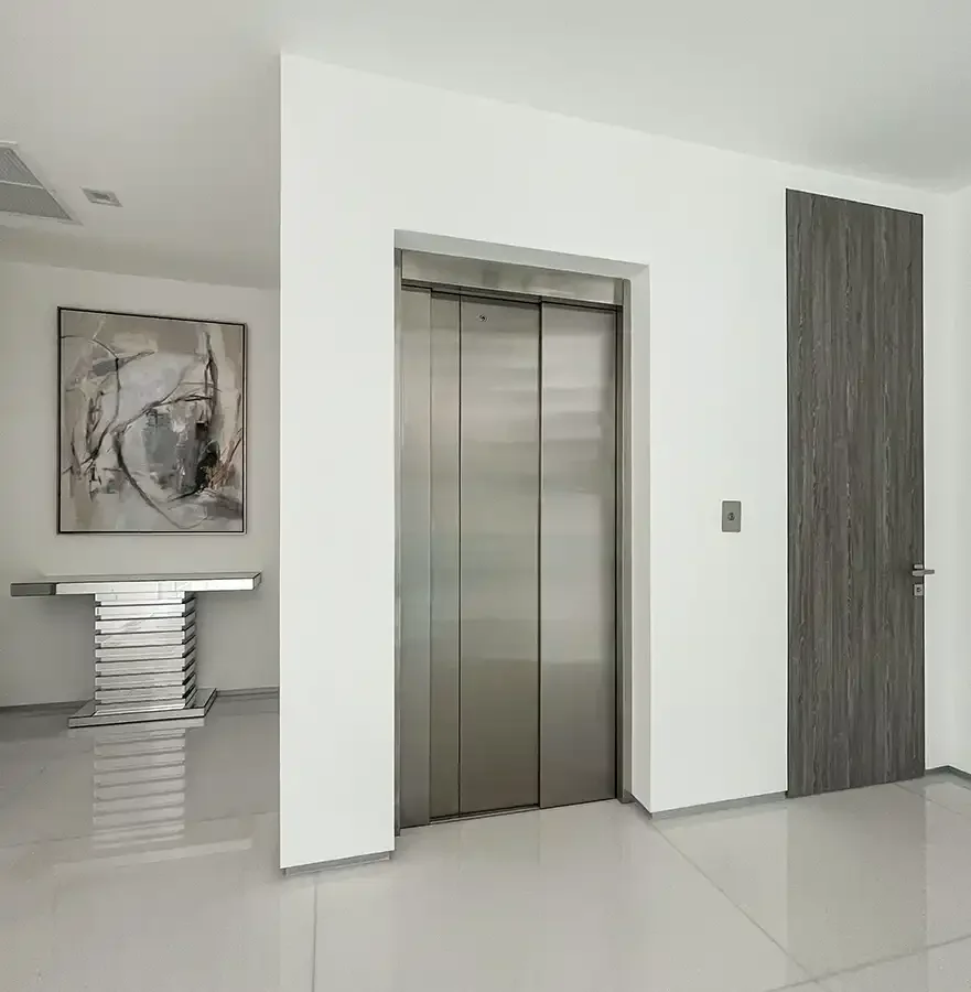 Residential elevator with stainless steel sliding doors installed in a minimalist modern interior, featuring white walls, polished flooring, and contemporary design.