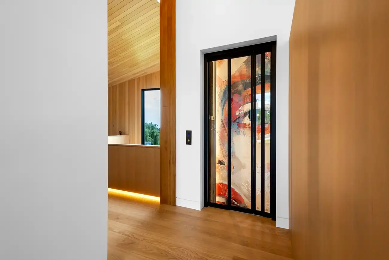 Residential elevator with custom art‑panel sliding doors and black frame, installed in a modern home interior with warm wood flooring and contemporary architectural finishes.