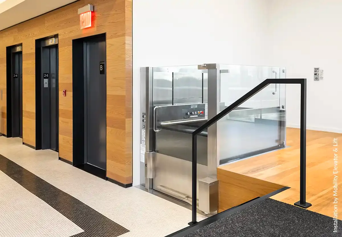 Glass-enclosed vertical platform lift adjacent to passenger elevators in a modern commercial lobby with wood paneling and glass railings