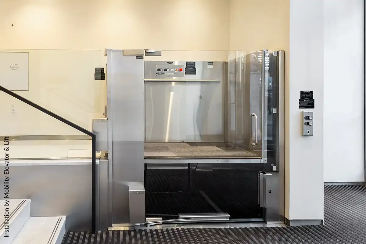 Open commercial vertical platform lift with stainless steel interior at a stair landing in a public building