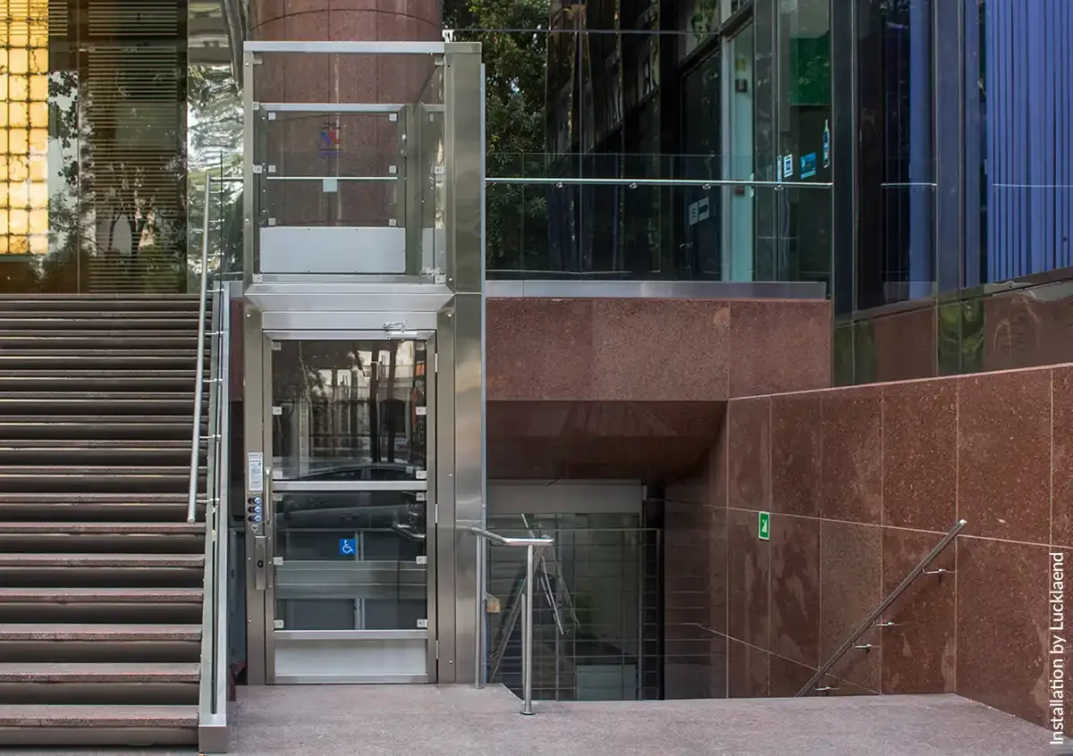 Glass-enclosed commercial platform lift adjacent to an exterior staircase at a modern building entrance