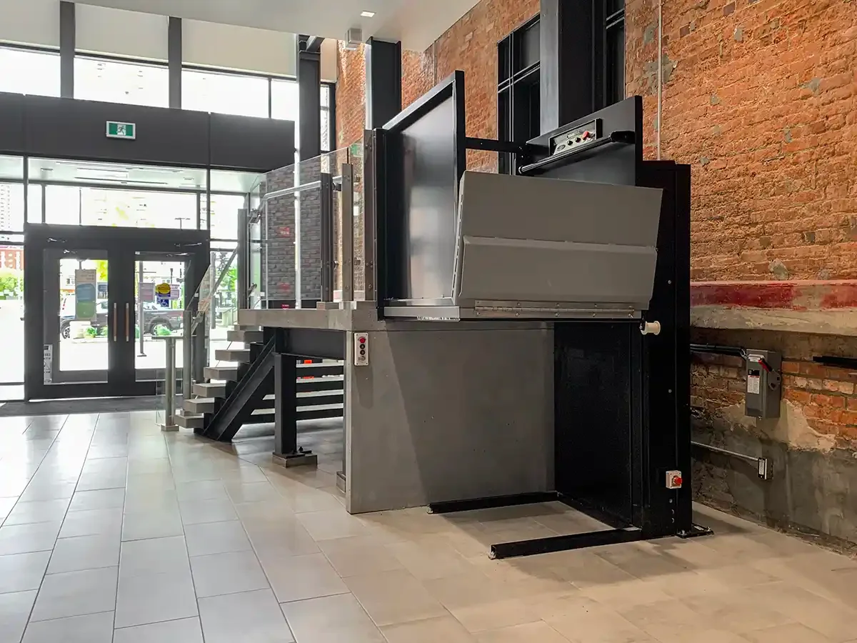 Indoor commercial vertical platform lift with black steel structure installed inside a brick building lobby, providing wheelchair accessibility between floor levels adjacent to stairs and glass entry doors.