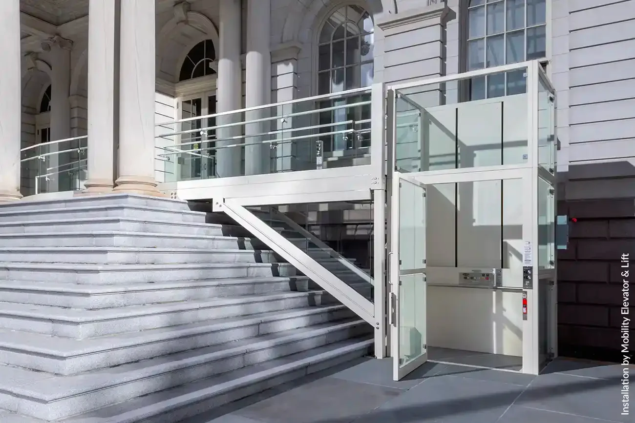 Glass-enclosed commercial wheelchair platform lift installed beside wide stone steps at a historic building entrance