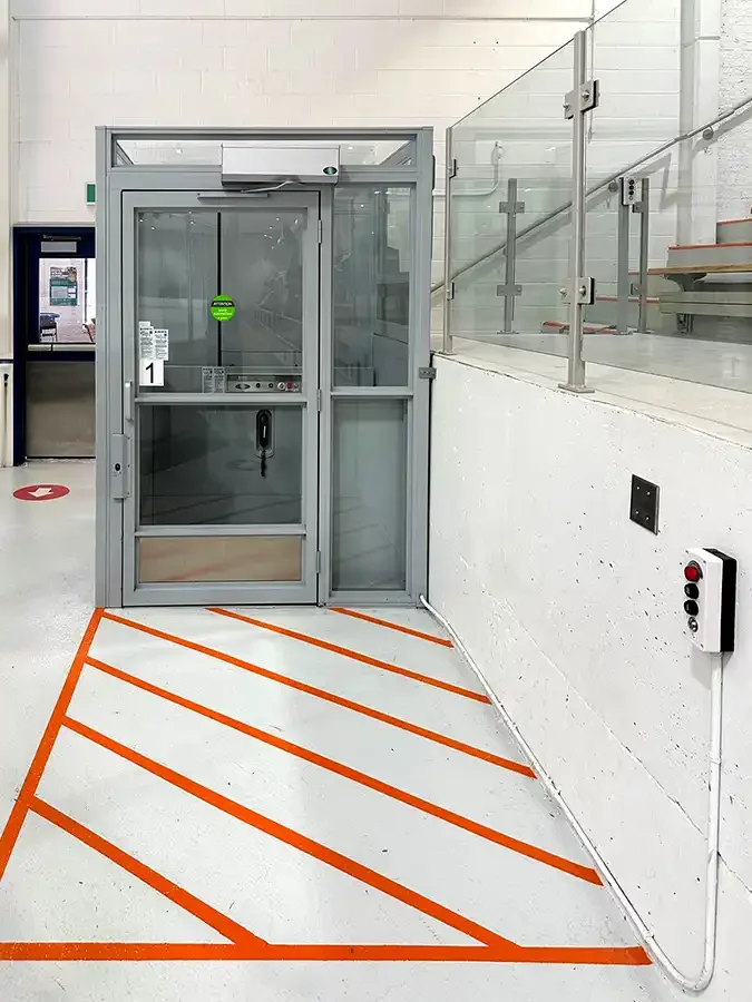 Glass-enclosed vertical platform lift in a commercial interior with floor safety markings and wall-mounted call controls