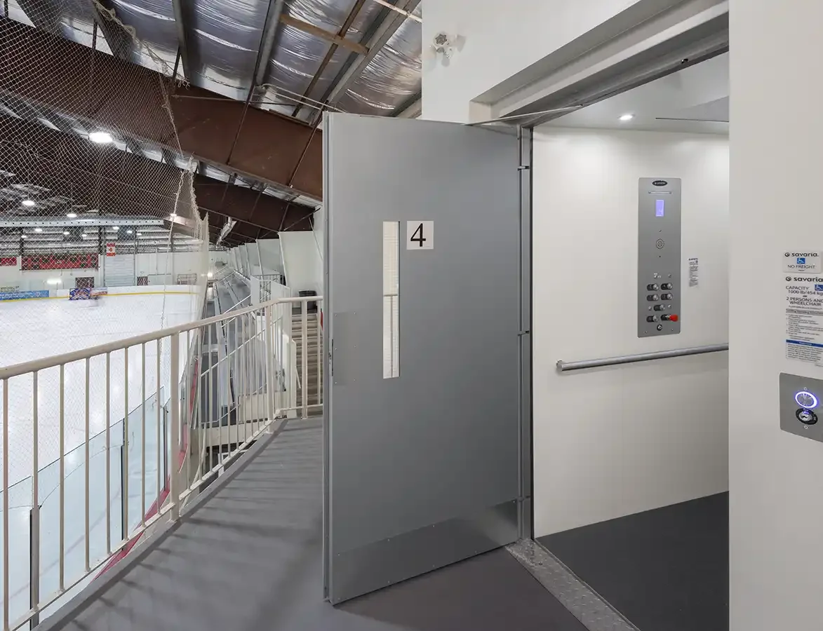 Open vertical platform lift behind a fire-rated commercial door overlooking an indoor ice rink and viewing area