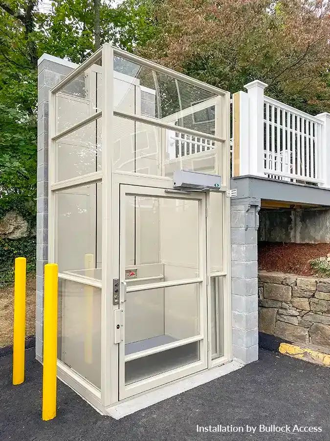 Glass-enclosed outdoor vertical platform lift installed beside a raised deck with white railing and stone foundation