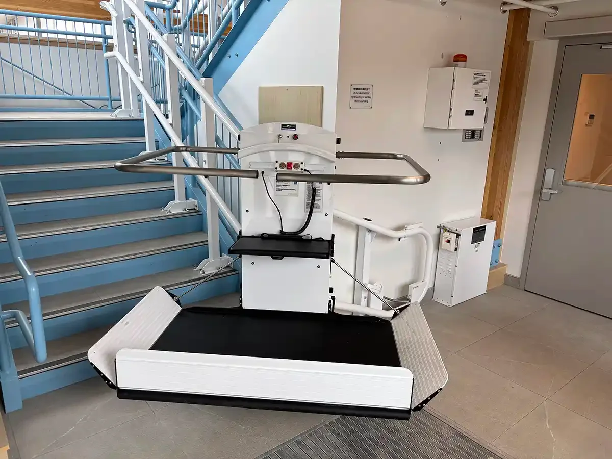 Indoor wheelchair platform lift positioned at the base of a commercial staircase with safety rails and folding platform