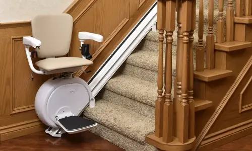 A Savaria K2 stationed at the bottom of a carpeted staircase with light brown wall moulding trim.