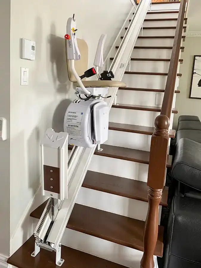 Stairlift with padded seat and armrests mounted on a straight rail along a wooden residential staircase with white risers and a wall-mounted control panel.