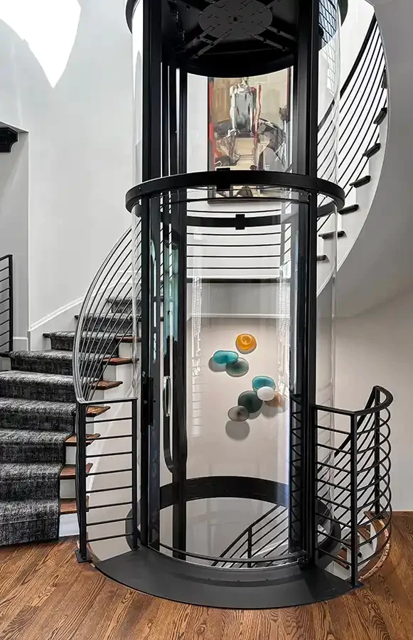Savaria Vuelift round panoramic home elevator with black powder-coated frame, glass panels, spiral staircase, and decorative wall art in contemporary entryway