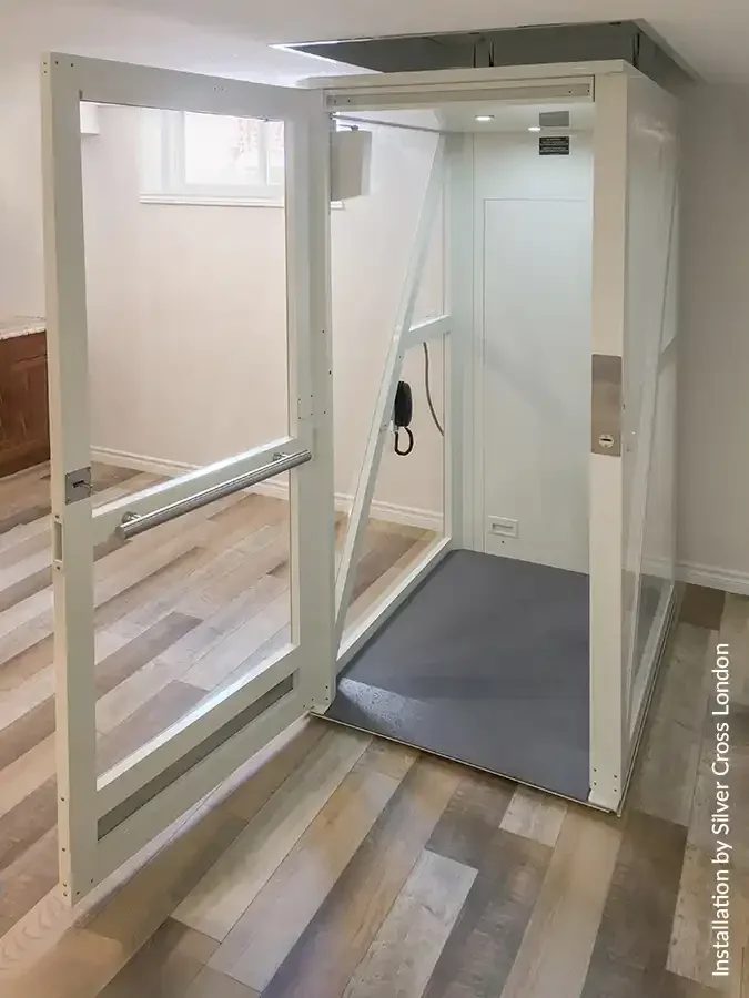 Through-floor home lift with open glass door and interior handrail positioned at floor level in a residential room with wood flooring.