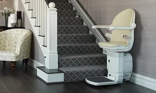 Stairlift parked at the bottom of a carpeted staircase, featuring a padded seat, armrests, and footrest mounted on a rail that runs along the side of the stairs in a traditional home interior.