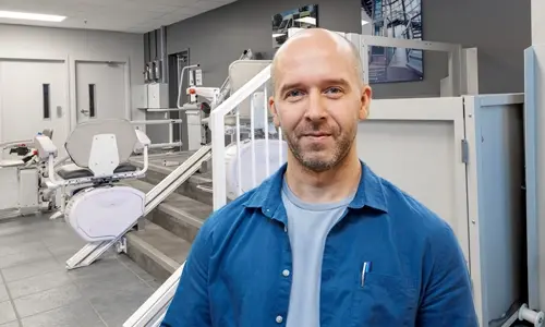 Person standing in a showroom-style space featuring multiple accessibility lifts, including stairlifts mounted on demonstration rails and a vertical platform lift beside a short staircase, with equipment and displays arranged throughout the room.