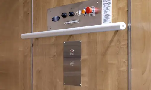 Stainless steel elevator control panel mounted on wood‑grain cab walls, featuring toggle switches, indicator lights, an emergency stop button, and a white horizontal handrail positioned below the controls.