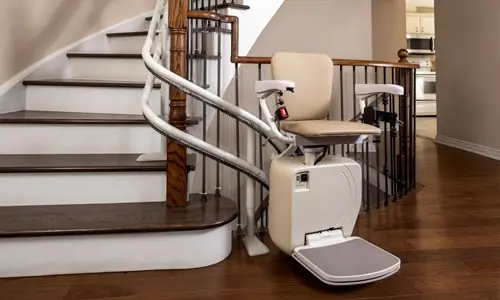 A curved stair lift installed along a wooden staircase, featuring a beige seat, footrest, and safety arms.
