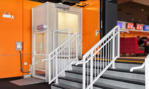 Vertical platform lift installed next to a bright orange wall in a bowling alley, providing accessible transport between levels.