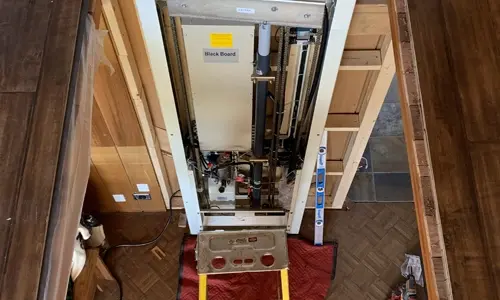 Residential home elevator machinery visible inside an open shaft, showing lift motor, rails, wiring, and mechanical components during installation or service.