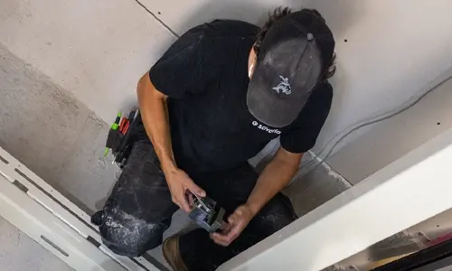 Elevator technician kneeling inside a residential home elevator shaft, installing metal hardware components during lift construction.