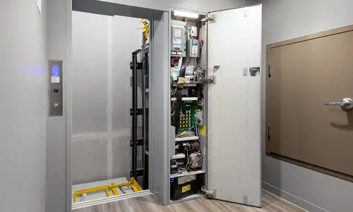 Residential home elevator with an open controller cabinet revealing electrical components, wiring, and lift machinery installed beside a finished hallway.