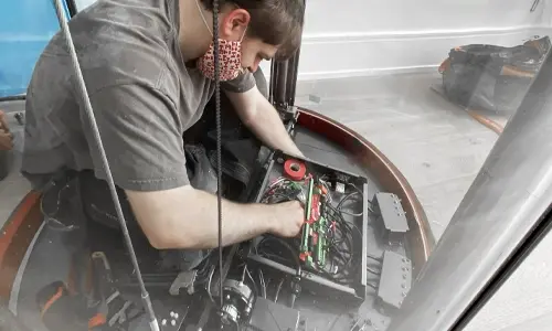 Elevator technician wearing a protective face mask performing electrical maintenance inside a panoramic glass residential elevator cabin.
